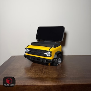 May include: A yellow and black 3D-printed phone stand shaped like the front end of an off-road vehicle. The stand features a black grille, circular headlights, and a storage compartment behind the phone mount. A black smartphone is docked in the stand.