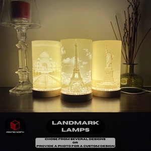 May include: Three illuminated cylindrical lamps with images of the Taj Mahal, the Eiffel Tower, and the Statue of Liberty. The lamps are on a wooden surface. The text "LANDMARK LAMPS" is below the lamps. The text "CHOSE FROM SEVERAL DESIGNS OR PROVIDE A PHOTO FOR A CUSTOM DESIGN" is below the text "LANDMARK LAMPS".