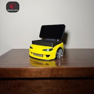 Puede incluir: Un soporte para teléfono con forma de coche amarillo impreso en 3D con un compartimento de almacenamiento negro y un teléfono negro. El coche tiene ruedas plateadas y un frontal detallado. El logotipo "PRINTED NORTH" está en la esquina superior izquierda.