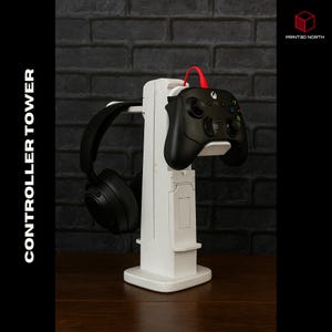 以下が含まれることがあります： 黒いXboxコントローラーと黒いヘッドフォンを備えた白いコントローラータワー。「CONTROLLER TOWER」の文字が側面にあります。赤いケーブルがコントローラーに接続されています。背景は暗いレンガの壁です。