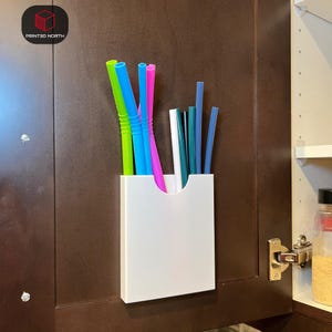 Puede incluir: Un recipiente de almacenamiento rectangular blanco con una abertura curva, montado en una puerta de armario marrón oscuro. El recipiente contiene una variedad de pajitas reutilizables de colores en verde, azul, rosa y blanco. El texto "PRINTED NORTH" es visible.