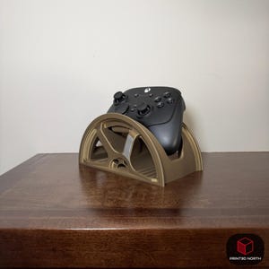 May include: A gold-coloured 3D-printed controller stand holding a black video game controller. The stand has a semi-circular design with a wheel-like pattern. The controller is resting in the stand, ready for use.