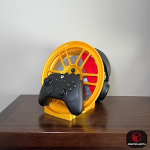 Könnte beinhalten: Ein gelber, 3D-gedruckter Gaming-Controller- und Headset-Ständer in Form eines Autorads. Ein schwarzer Xbox-Controller liegt vorne, mit einem schwarzen Headset und roten Akzenten hinter dem Rad. Der Ständer steht auf einer dunkelbraunen Oberfläche.
