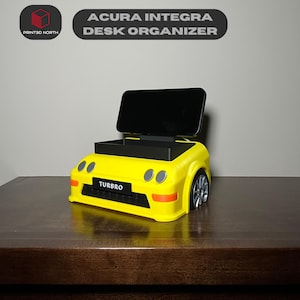 May include: Yellow 3D printed Acura Integra desk organiser with a phone stand and a compartment for storing small items. The organiser has a "TURBRO" label on the front.