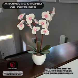 May include: A white ceramic orchid plant diffuser with pink flowers. The diffuser is designed to hold essential oils or incense sticks. The text "A GORGEOUS ORCHID 'PLANT' FOR DIFFUSER OILS OR INCENSE STICKS" is printed on a white label below the diffuser.