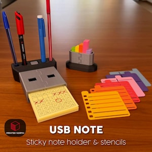 May include: A black USB-shaped sticky note holder with pens and a notepad. The notepad shows a calendar with dates marked. Also included are colourful stencils and a small organiser with colourful blocks. The text "USB NOTE" is visible.