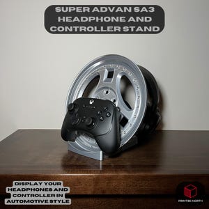 May include: Silver headphone and controller stand with the text "SUPER ADVAN SA3 HEADPHONE AND CONTROLLER STAND". A black controller rests on the stand, with a pair of headphones behind it. The stand is designed to display headphones and controllers in automotive style.