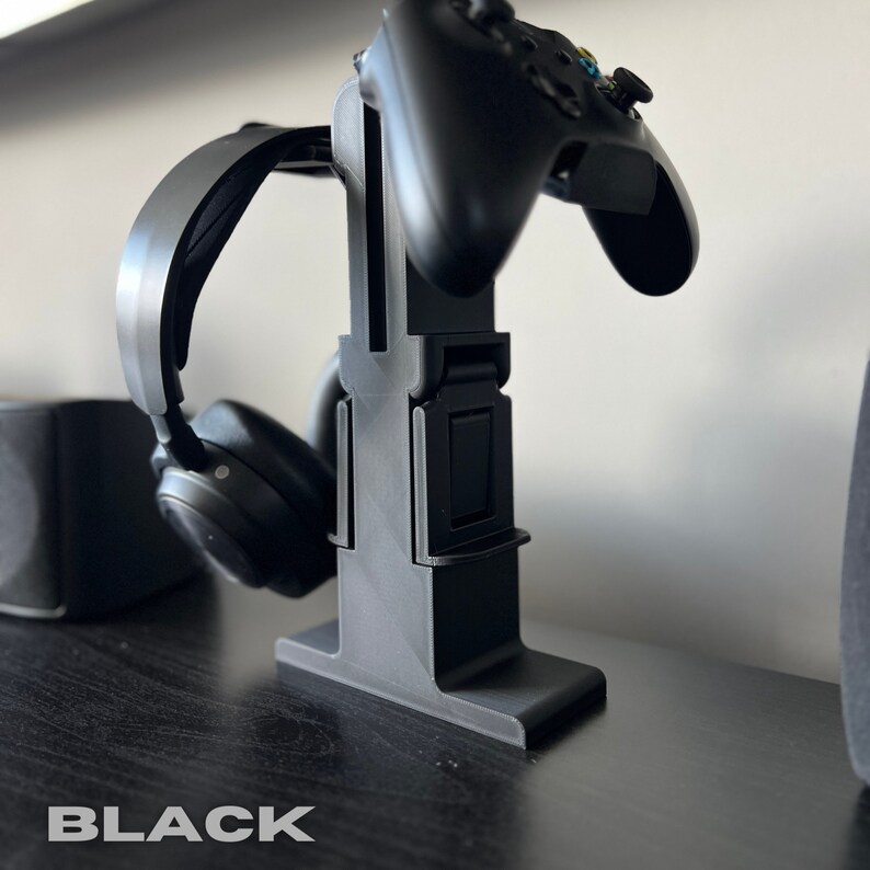 May include: Black gaming headset stand with a controller holder. The stand is made of plastic and has a sleek, modern design.