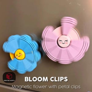 Puede incluir: Dos clips magnéticos con forma de flor, uno azul y otro rosa, con caras sonrientes en el centro. El clip azul tiene un centro amarillo, mientras que el clip rosa tiene un centro blanco. El texto "BLOOM CLIPS" y "Magnetic flower with petal clips" son visibles.