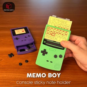 Peut inclure: Un porte-notes adhésives en forme de console portable, disponible en violet et en vert lime. Le modèle vert contient une note jaune avec un calendrier de septembre marqué de croix rouges. Le modèle violet possède un tiroir avec deux objets noirs.