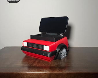 May include: A red 3D printed car phone holder with a black phone stand and a small storage compartment. The car is a Volkswagen Golf.