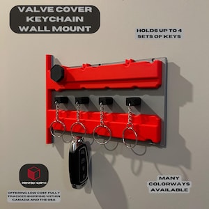 Puede incluir: Porta llaves de pared impreso en 3D de color rojo y gris con forma de tapa de válvula. Tiene cuatro llaveros y el texto "HOLDS UP TO 4 SETS OF KEYS" y "MANY COLORWAYS AVAILABLE".