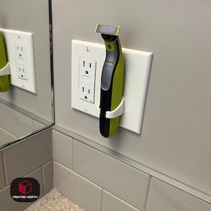 Puede incluir: Una afeitadora eléctrica Philips OneBlade negra y verde se sujeta en un soporte blanco impreso en 3D, fijado a una cubierta de enchufe blanca. La afeitadora está colocada verticalmente. La pared es de color gris claro.