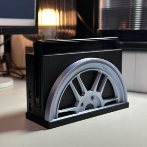 Peut inclure: Un support d'impression 3D noir pour une console Nintendo Switch, avec un design de roue argenté.