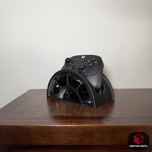 May include: Black Xbox controller resting on a black 3D-printed controller stand. The stand has a semi-circular design with a series of circular cutouts. The controller is centered on the stand, showcasing its buttons and joysticks.