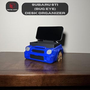 May include: Blue Subaru STI "Bug Eye" car-shaped desk organiser with a phone stand. The organiser has a compartment for storing small items.