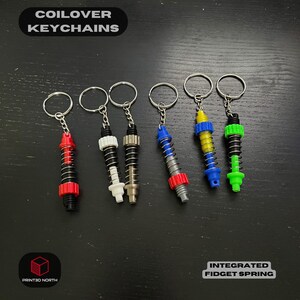 May include: A collection of coilover keychains in various colors, including red, white, black, blue, yellow, and green. Each keychain features a miniature spring design with a metal ring for attaching keys. The text "COILOVER KEYCHAINS" and "INTEGRATED FIDGET SPRING" are visible.