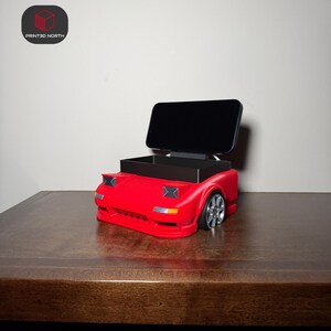 Peut inclure: Un support de téléphone rouge imprimé en 3D en forme de voiture avec un téléphone noir en place. Le support a un compartiment de rangement et des phares et des roues détaillés. Le logo "PRINTED NORTH" est dans le coin supérieur gauche.