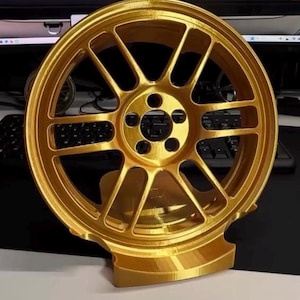 May include: A gold-coloured car wheel with multiple spokes and a central hub with five holes. The wheel is displayed on a stand. The background includes a car and the text "GTI".