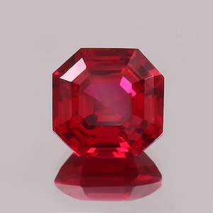 May include: A faceted, square-cut ruby gemstone. The gem is a vibrant red color with a reflective surface, and it is set against a light background. The gemstone's facets create a play of light and shadow, enhancing its brilliance.