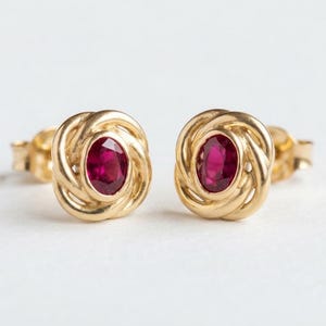 Pode incluir: Um par de brincos de ouro com uma pedra preciosa de rubi vermelho escuro de corte oval no centro, rodeada por uma moldura de metal dourado polido com um padrão de nó entrelaçado. Os brincos incluem fechos de pino para fixação segura.