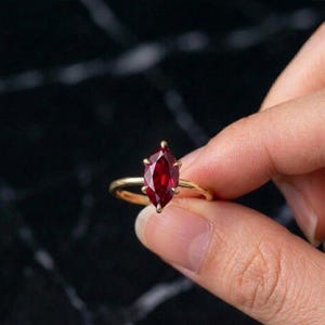 May include: A gold ring with a marquise-cut, deep red gemstone. The ring's band is thin, polished gold, and the gemstone is held by delicate prongs. The ring is shown against a dark, textured background.