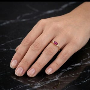 Könnte beinhalten: Ein goldener Ring mit einem rechteckigen, tiefroten Edelstein als Hauptmerkmal. Kleine, klare Steine ​​sind auf beiden Seiten des roten Steins angeordnet. Der Ring wird auf einer Hand vor einem dunklen, strukturierten Hintergrund gezeigt.