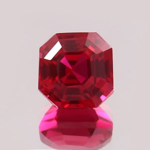 May include: A faceted, square-cut ruby gemstone. The gem is a deep, vibrant red color with a high degree of clarity. The facets reflect light, creating a brilliant sparkle. The gemstone is set against a white background, with its reflection visible below.