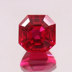 May include: A faceted, octagonal ruby gemstone. The gem is a vibrant, deep red color with a high degree of clarity. The facets reflect light, creating a brilliant sparkle. The gemstone is set against a white background, with its reflection visible below.