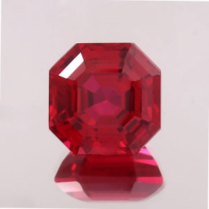May include: A faceted, octagonal-cut ruby gemstone. The gem is a deep, saturated red color, with sharp, clean facets that reflect light. The gemstone is set against a white background, with its reflection visible below.
