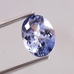 Op de afbeelding: Een lichtblauwe, ovaalvormige edelsteen die door een metalen pincet wordt vastgehouden. De edelsteen heeft een gepolijst oppervlak met meerdere facetten, dat licht reflecteert. De edelsteen is lichtblauw.