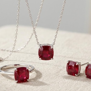 Könnte beinhalten: Ein Schmuckset mit einer silbernen Halskette, einem Armband, einem Ring und Ohrsteckern. Jedes Schmuckstück zeigt einen quadratischen, tiefroten Edelstein. Der Schmuck wird auf einer neutralen Oberfläche präsentiert.