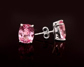 May include: A pair of stud earrings featuring oval-cut, light pink gemstones set in a silver metal. The earrings are displayed against a dark background, highlighting their color and design. The gemstones have a faceted appearance.