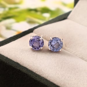 May include: A pair of round, light purple gemstone stud earrings. Each earring features a faceted, circular stone held in place by four silver prongs. The earrings are displayed in a dark green jewelry box with a light grey interior.