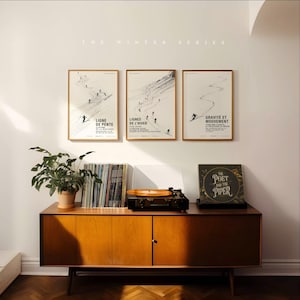 May include: Three framed art prints from "The Winter Series" featuring skiers on slopes, with French text. A mid-century modern wooden cabinet holds a record player, vinyl records, and a potted plant. The album cover reads "The Poet and the Piper."