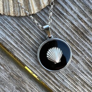 Op de afbeelding: Een zilveren ketting met een ronde hanger met een zwarte achtergrond en een zilveren schelpontwerp.