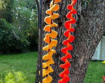 May include: Two crocheted yarn spirals hanging from a black metal stand. The spiral on the left is yellow and orange, while the spiral on the right is red and orange.