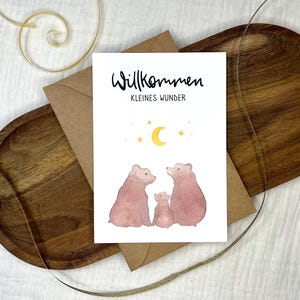 Könnte beinhalten: Eine weiße Karte mit dem Text "Willkommen Kleines Wunder" in schwarzer Schrift. Die Karte zeigt eine Aquarell-Illustration von zwei braunen Bären und einem kleineren Bärenjungen, die zu einem gelben Halbmond und Sternen aufblicken.