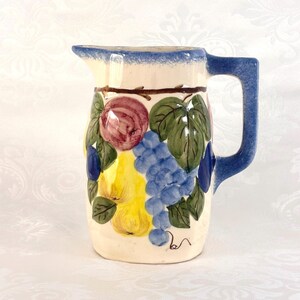 May include: A ceramic pitcher with a blue handle and rim, decorated with painted fruit and leaves. The fruit includes grapes, pears, and a red-toned fruit. The pitcher has a cream-colored base.