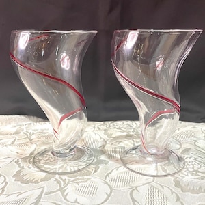 May include: Two clear glass drinking glasses with a red swirl design. The glasses have a unique shape with a curved base and a wide, open top.
