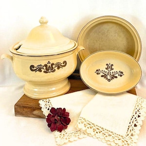 May include: A cream-colored ceramic soup tureen with a lid, a serving plate, and an oval dish, all featuring a brown floral design. Two white lace-edged napkins and a burgundy flower complete the arrangement. The set is displayed on a wooden surface.