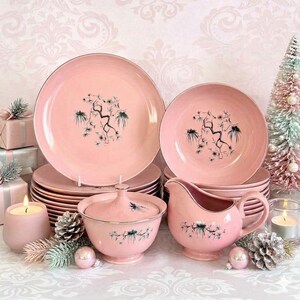 May include: A set of pink ceramic dinnerware featuring a dark green pine branch illustration. The collection includes stacked dinner plates, salad plates, a lidded sugar bowl, and a gravy boat, all decorated with a consistent pine motif.