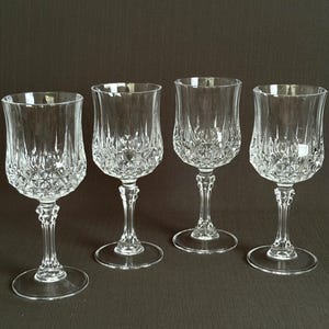 May include: Four clear crystal wine glasses with diamond-cut patterns on the bowls. The glasses have long, slender stems and round bases. The background is a solid dark brown.