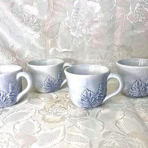 May include: Four ceramic mugs with a light blue glaze and leaf design. Each mug has a handle and a leaf motif on the side. The mugs are arranged on a white, textured surface.