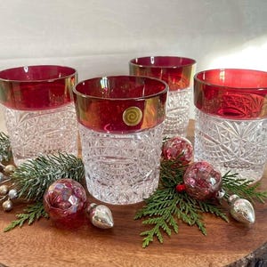 May include: A set of four clear glass tumblers featuring a cut crystal pattern on the lower half and a smooth, deep red band around the top rim. Each glass has a small, circular gold-colored sticker with a spiral design on the red rim.