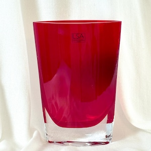 May include: A vibrant, ruby-red glass vase with a clear, rounded base. The vase has a rectangular shape with a slightly curved bottom. The LSA brand logo is visible on the front. This decorative vase is suitable for flowers or as a standalone art piece.