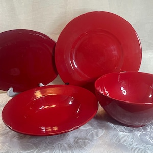 May include: A set of four glossy red ceramic dinnerware pieces including one large round dinner plate, one oval serving platter, one shallow wide-rimmed bowl, and one deep round bowl, all featuring a smooth, solid, and reflective finish.