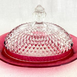 May include: A clear glass butter dish with a ruby red base and lid. The lid is domed with a diamond pattern and a small, decorative handle. The base is a solid, round plate. The dish is designed for serving butter.