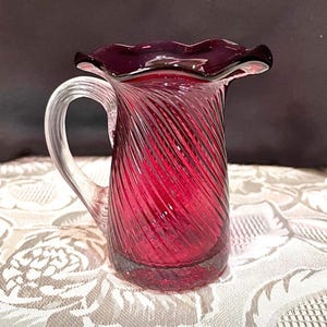 May include: A ruby red glass pitcher with a clear handle. The pitcher has a ruffled rim and a spiral design. The handle is clear glass. The pitcher is likely used for serving beverages.