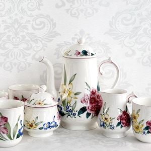 May include: A white floral tea set featuring a teapot, creamer, sugar bowl, and cups. The set is decorated with colorful flowers in shades of pink, yellow, and blue, with green leaves. The rims of the cups and teapot are trimmed in a burgundy color.